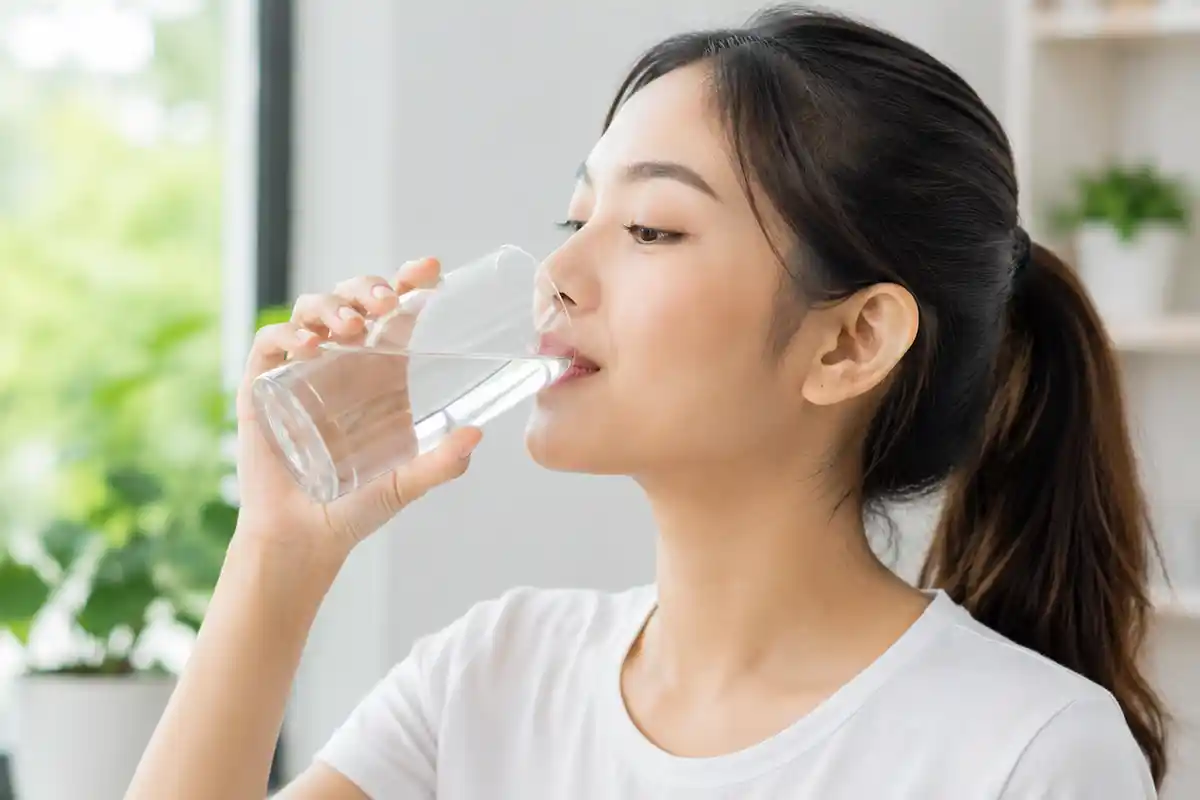 Seorang wanita sedang minum segelas air putih segar dengan latar belakang ruangan rumah yang terang dan tanaman hijau.