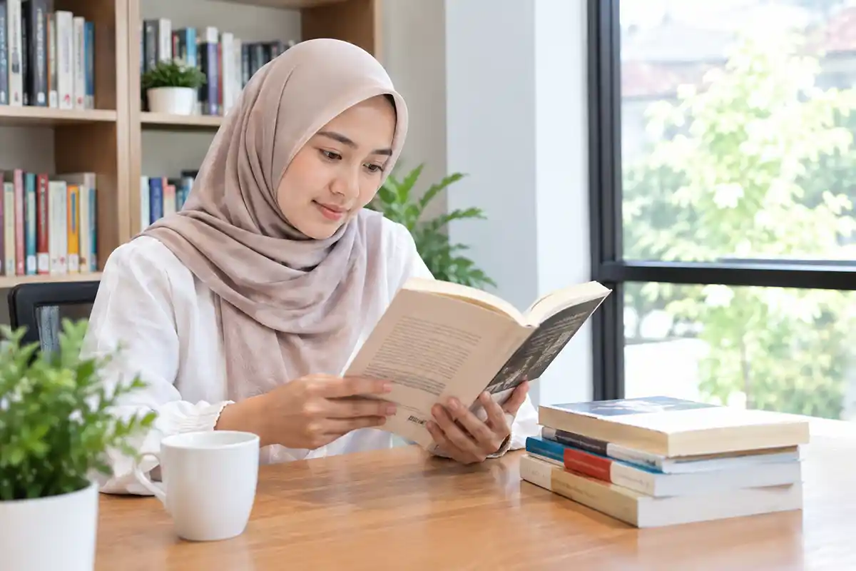 Seorang wanita berhijab sedang fokus membaca buku di meja dekat tumpukan buku.