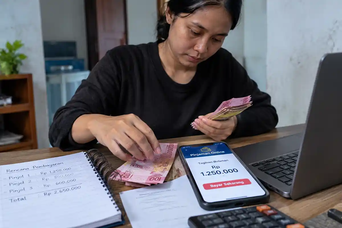 Seorang wanita sedang menghitung uang tunai di meja kerja sambil melihat tagihan pinjaman online di HP.