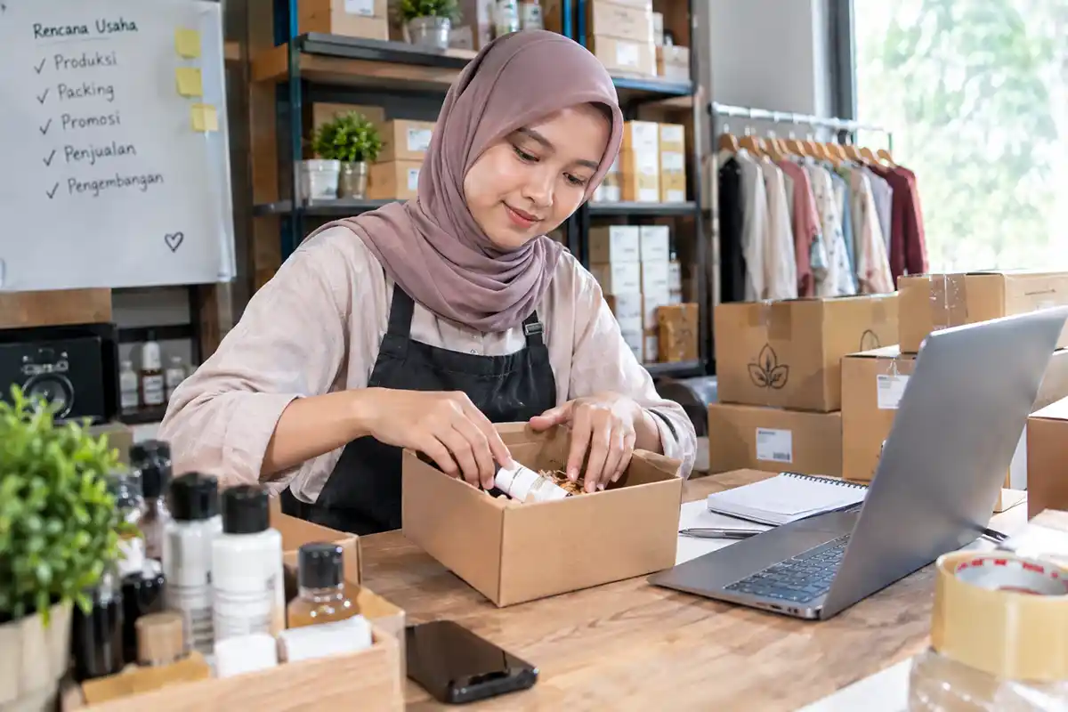 Pelaku usaha wanita berhijab sedang mengemas produk ke dalam kardus di meja kerja berlaptop.