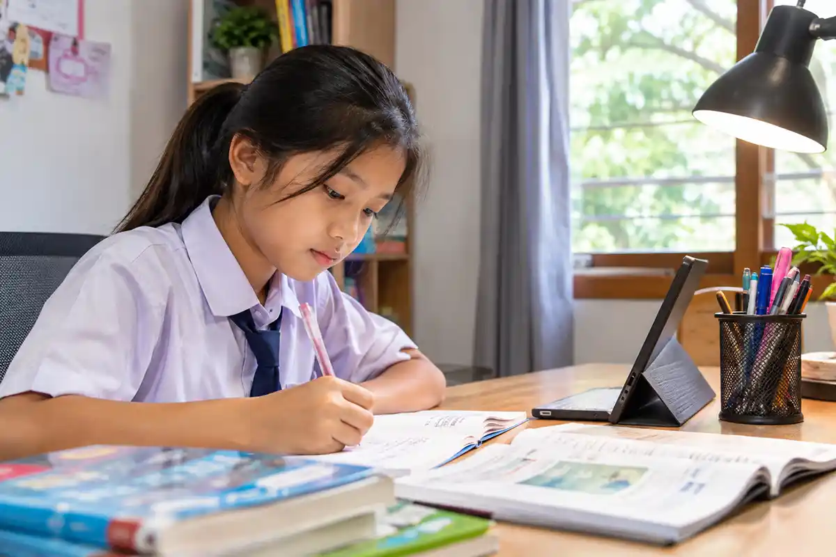 Seorang anak perempuan berseragam sekolah sedang fokus belajar dan menulis di meja belajar.