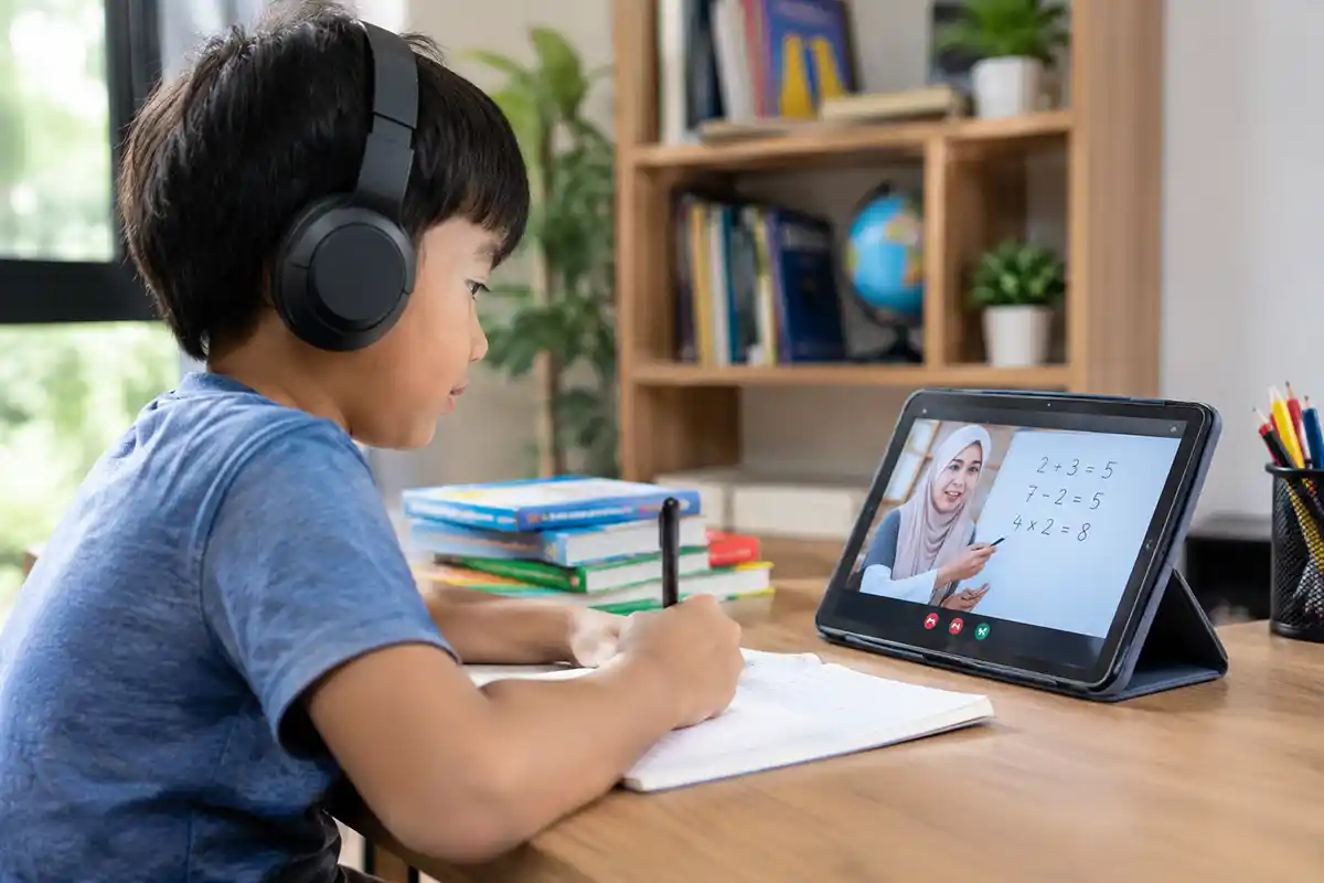 Seorang anak sekolah memakai headphone sedang menyimak sesi belajar daring dengan guru melalui tablet.
