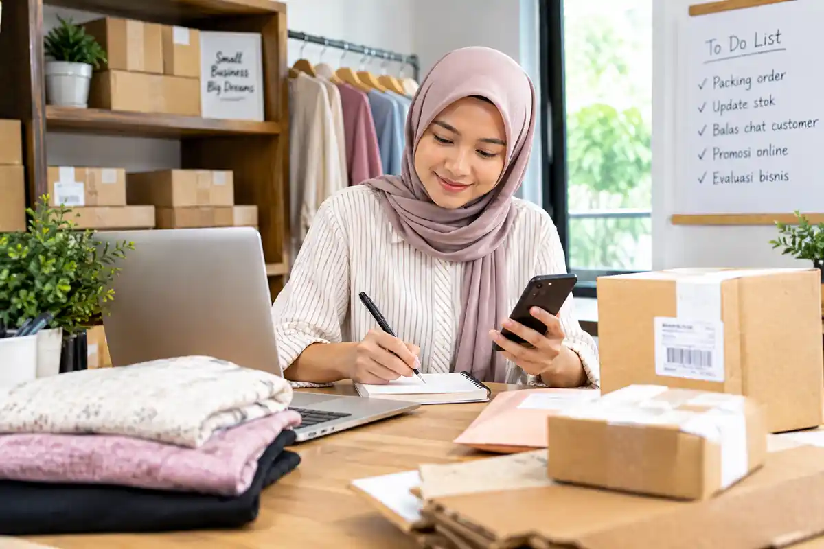 Wirausahawan wanita berhijab mengelola pesanan toko online dan mencatat daftar tugas di meja kerja.